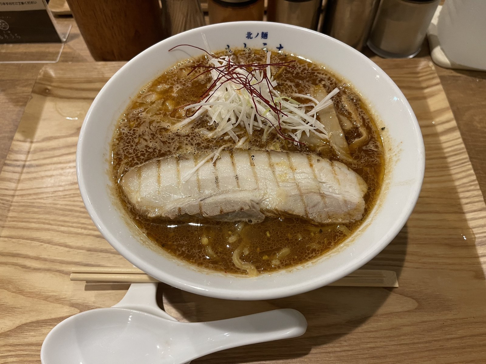 札幌スパイシーラーメンこくうま中辛の全体、糸唐辛子と白髪ねぎがのった茶色いスープに大きなバラチャーシューが浮かぶ
