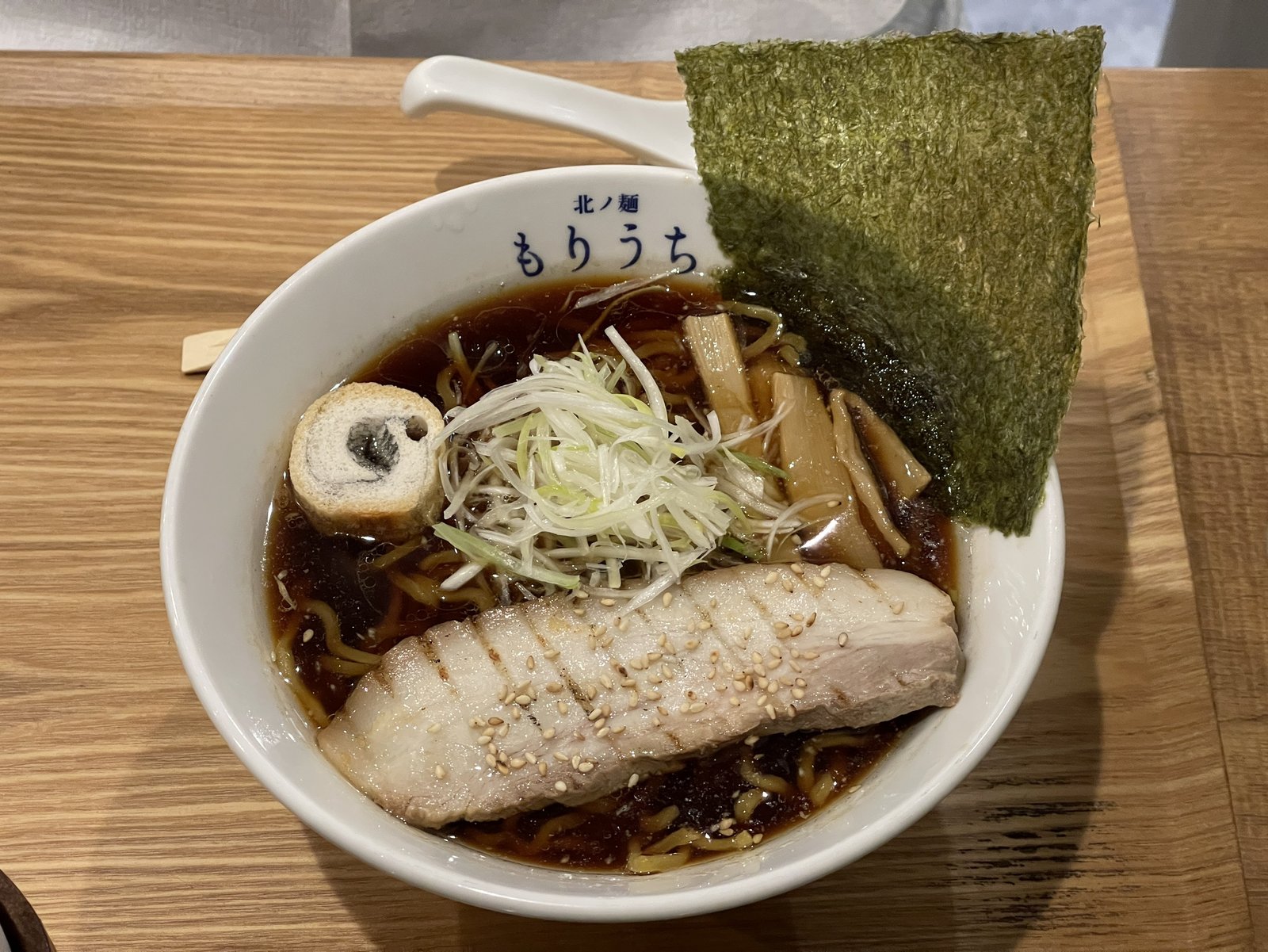 黒醤油ラーメンの全体、濃い色のスープに大きな海苔とお麩、白髪ねぎ、メンマ、バラチャーシューがのっている
